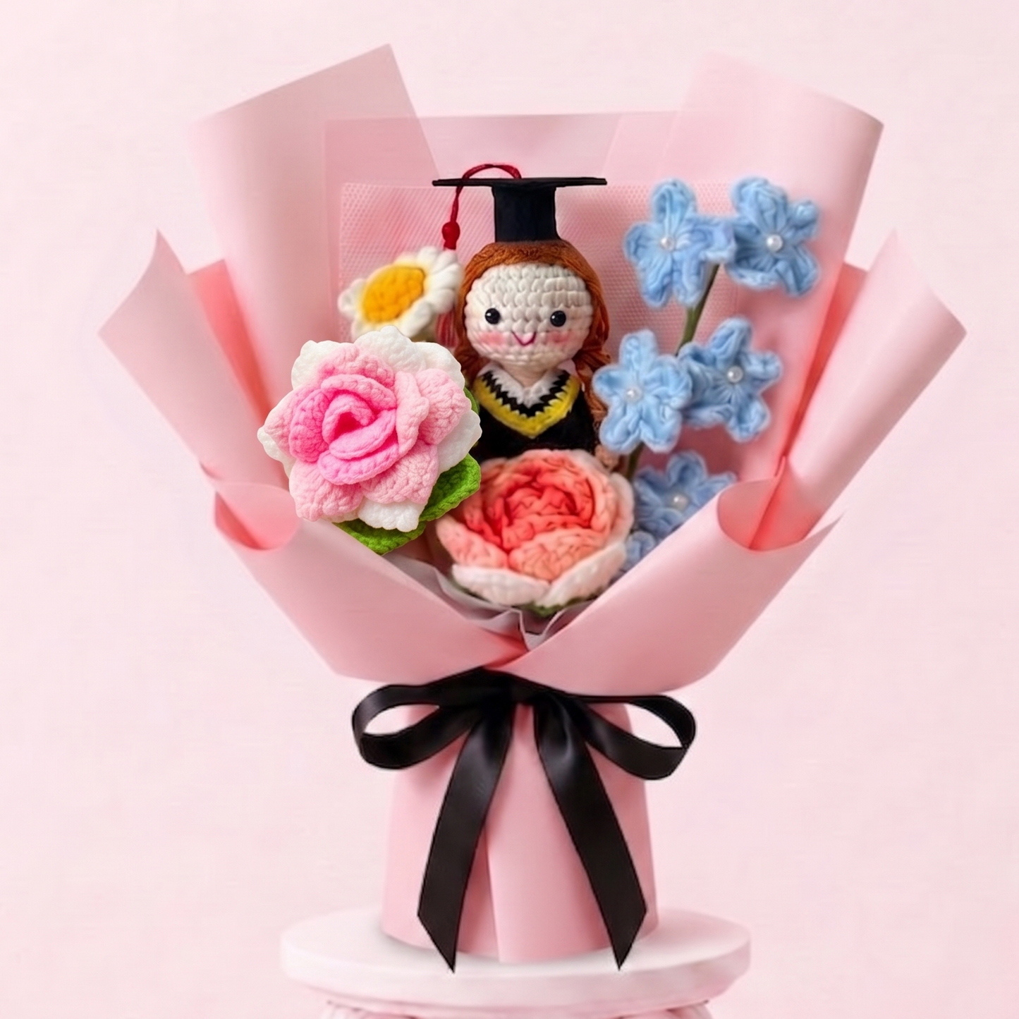 A handmade crochet flower bouquet gift set on a pink pedestal. The bouquet features pink and red roses, blue flowers, a small crochet bee, and a graduation cap, wrapped in pink paper with a black ribbon bow.