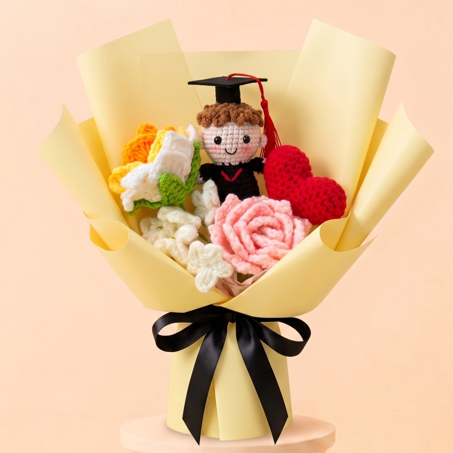 A handmade crochet graduation bouquet wrapped in pale yellow paper, featuring a crocheted graduate figure in a black cap and gown, pink and red roses, white flowers, and green leaves, displayed on a decorative pedestal against a soft peach background.